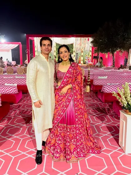 I love designing for couples, helping them coordinate for special events. Here, my husband wears a custom-made beige kurta pajama with subtle leatherwork, while I am in a vibrant pink patola lehenga paired with a hand-embroidered gota blouse.