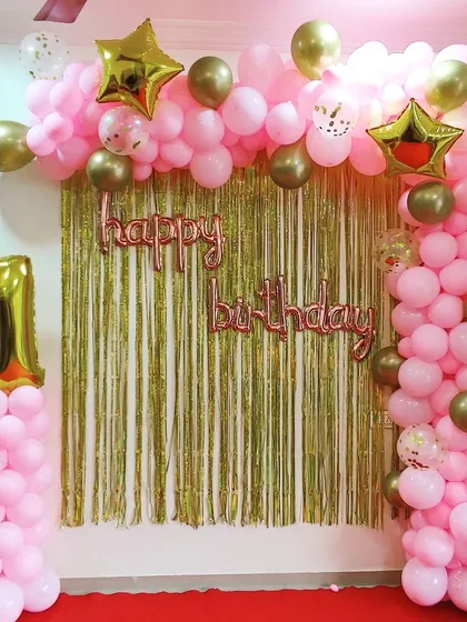 A pretty-in-pink first birthday decoration. The setup features a pink and gold balloon arch against a gold shimmer curtain, with a large '1' foil number on a matching balloon pillar.