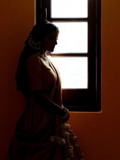 A silhouette of the bride, Deeksha, standing by a window. The warm light and her quiet pose create a moment of solitude and contemplation before her haldi ceremony.