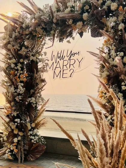 A closer look at the "Will you MARRY ME?" neon sign against the sunset. The sign is integrated into the pampas and floral arch, ensuring it’s a beautiful focal point for photos.