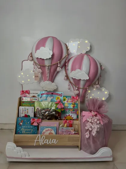Up, up, and away! This hot air balloon themed gift for a little girl named Alaia features a personalized wooden bookshelf filled with storybooks, all under a dreamy sky of balloon models and glowing clouds.