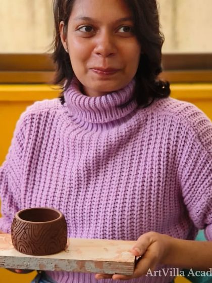 A student proudly shows off her hand-built pot with a carved texture. The smile on her face reflects the satisfaction of creating something beautiful with her own hands.