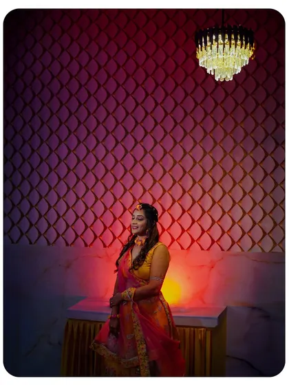 A wide shot that captures the ambiance of the Haldi ceremony. The bride is framed against a textured wall under a chandelier, with dramatic lighting that creates a moody and beautiful portrait.
