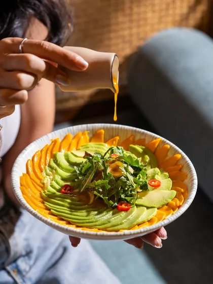 A person drizzling dressing over a fresh mango and avocado salad. The human element makes the photo more dynamic and relatable.