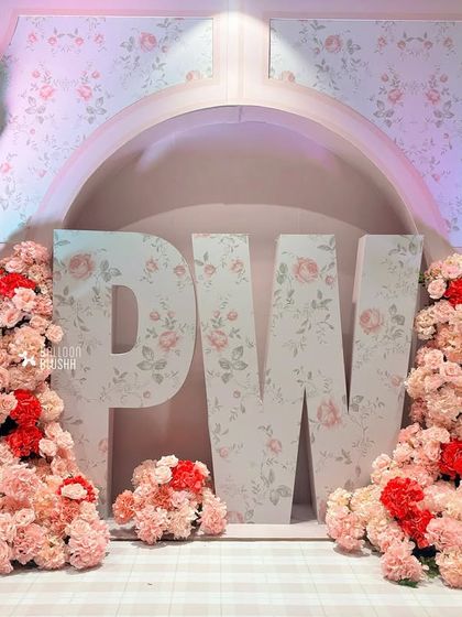 A head-on shot of the "PW" floral installation. The combination of floral-print letters and dense clusters of pink and red carnations creates a lush, textured, and highly photogenic backdrop.
