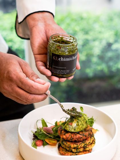 Our Chimichurri is a versatile sauce that can elevate any dish. Here, a chef spoons it over a stack of vegetable fritters, adding a fresh, herby kick.