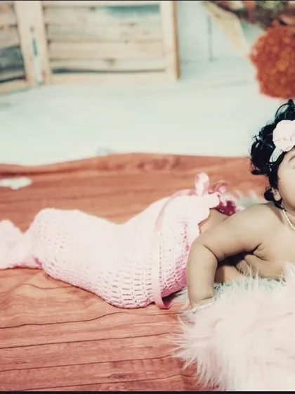 An adorable baby girl dressed as a little mermaid in a pink crocheted outfit, lying on a soft rug.