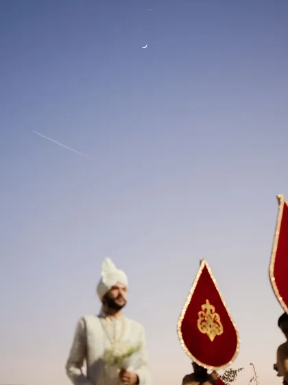 An artistic shot of the groom's procession, with the crescent moon and a jet stream in the sky above. This photo captures a unique and almost poetic moment during the wedding festivities.
