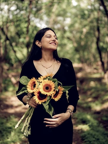 A joyful portrait of the mom-to-be in a sunlit forest, holding a large bouquet of sunflowers and smiling with pure happiness.