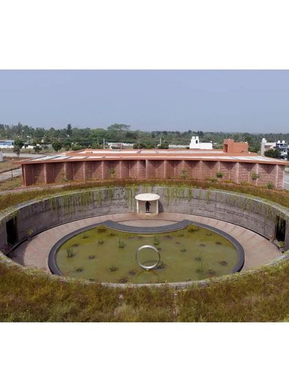 A wider perspective of the memorial, showing the sunken circular gallery and the larger auditorium building behind it. The design uses changes in elevation to create distinct zones for quiet reflection and active community use, all unified by a consistent architectural language.