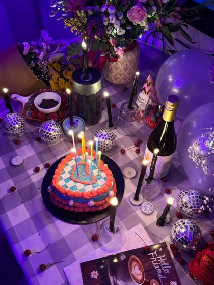 A vibrant, heart-shaped birthday cake with a colorful checkered pattern, set amidst a fun, candlelit party scene with disco balls and balloons.