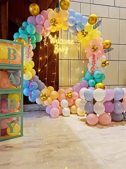 A beautiful pastel-themed welcome home decoration. This setup features a full circular balloon arch in rainbow colors, a "Welcome Baby" neon sign, and matching "BABY" display blocks.