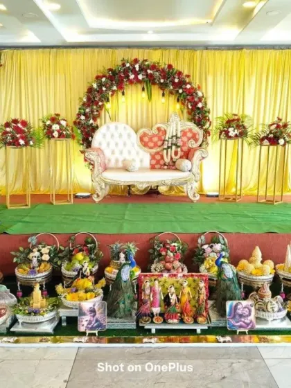 A wide-angle view of a traditional ceremony stage. The setup includes a red rose arch, a beautiful diwan, and a detailed arrangement of dolls and gift platters, creating a festive atmosphere.