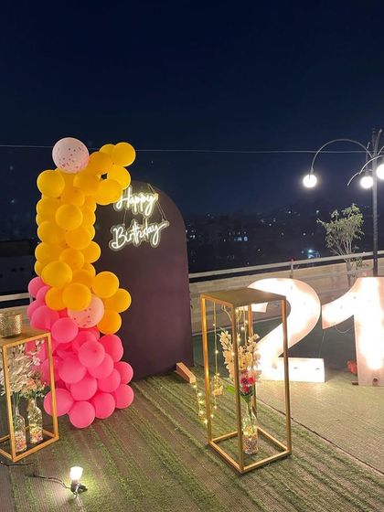 A vibrant 21st birthday decoration on a rooftop. This setup features a balloon arrangement in hot pink and yellow, a 'Happy Birthday' neon sign, and large marquee numbers against a city skyline.