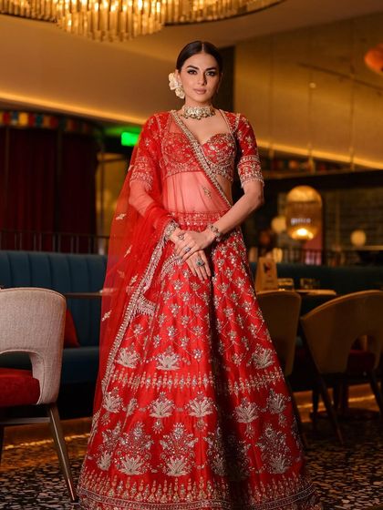 A royal wedding look featuring a classic red lehenga. The makeup is clean and defined, with a sleek bun adorned with flowers, creating a look of timeless bridal elegance.