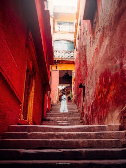 The narrow, ancient alleyways of Varanasi, painted in vibrant red. A lone figure in white stands on the steps, creating a powerful contrast and a sense of timelessness.