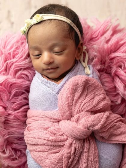 A tiny newborn baby wrapped in soft pink and purple fabrics, nestled in a fluffy basket with a little bunny friend.