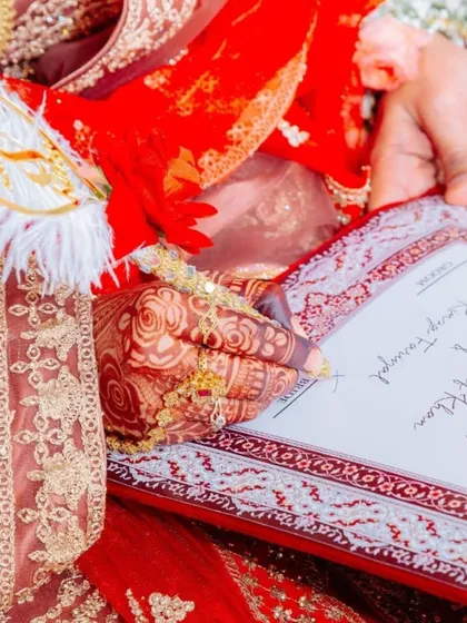 A close-up shot of the bride's henna-adorned hand as she signs the Nikah nama. The decorated pen and the intricate border of the certificate add to the beauty of the moment.