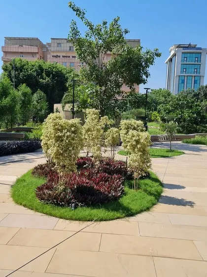 A central planting feature in a public park, with a variety of shrubs creating a textured, multi-colored island. This serves as a focal point along the paved walkway, breaking up the expanse of the path.