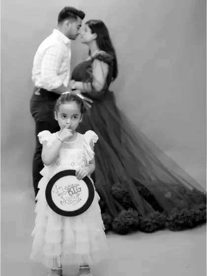 A creative family maternity photo announcing the arrival of a new sibling. The older sister takes center stage while her parents share a moment in the background.