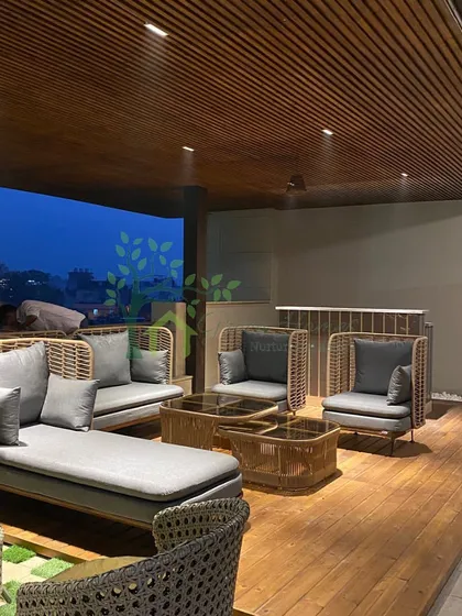 A real photograph of a completed rooftop project in Delhi. This image shows the modern outdoor furniture on a wooden deck under a covered pergola, overlooking the city at twilight.