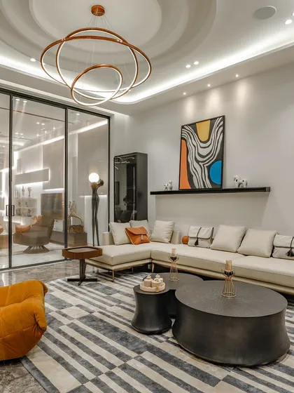 A wide shot of a contemporary living room, featuring a large sectional sofa, a vibrant orange lounge chair, and a modern circular chandelier. The glass partition offers a glimpse into an adjacent space, enhancing the open feel.