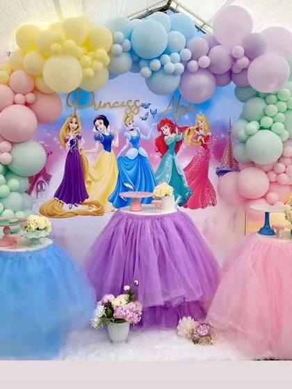 A beautiful pastel princess party setup with tutu-covered tables. The backdrop features classic princesses under a soft, cloud-like balloon arch.