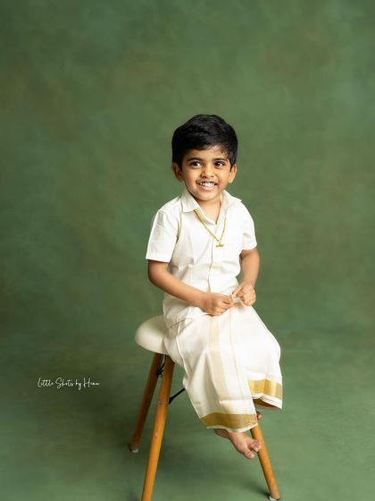 A handsome little boy dressed in a traditional veshti and shirt. This simple, classic portrait against a solid green backdrop highlights his bright smile and cultural attire.