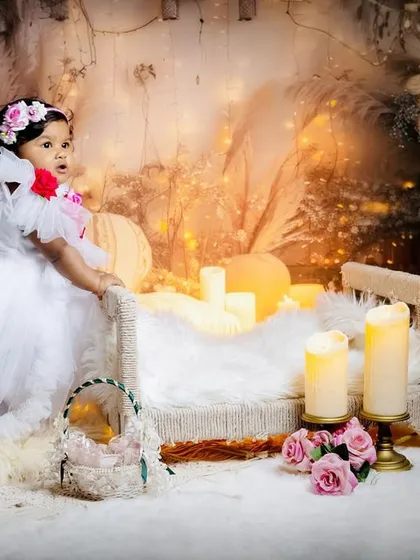 A baby girl in a white dress stands by a miniature bed in a dreamy, candle-lit boho setup for her sitter session.