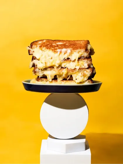A towering stack of gooey grilled cheese sandwiches, balanced on geometric blocks against a bold yellow background. This quirky and minimalist shot is designed to be eye-catching and memorable.