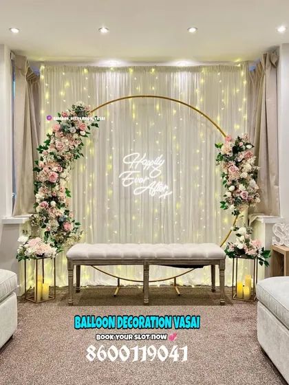 For an elegant engagement or anniversary party, I can create a beautiful photo backdrop like this one. It features a large golden ring decorated with soft pink and white floral arrangements, set against a backdrop of twinkling fairy lights and a "Happily Ever After" neon sign.