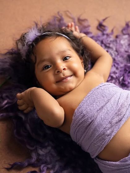 A happy six-month-old lies on a fluffy purple rug, smiling brightly for the camera. Milestone sessions are perfect for capturing this joyful age.