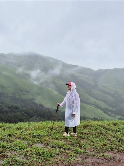 A lone trekker in a poncho stands amidst the green meadows of Netravathi, taking in the misty landscape.