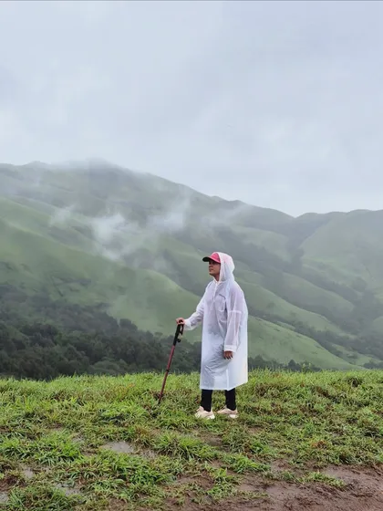 A lone trekker in a poncho stands amidst the green meadows of Netravathi, taking in the misty landscape.
