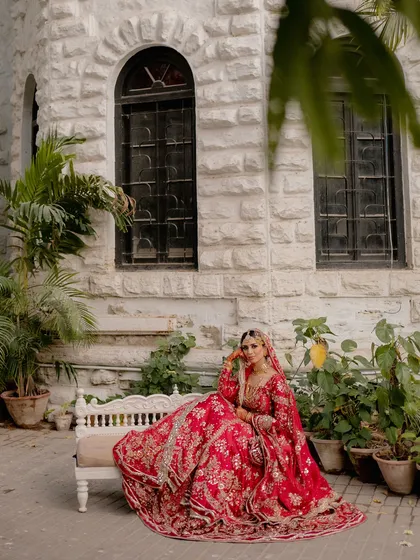 A moment of quiet contemplation for the bride. Seated gracefully, her vibrant red lehenga spread around her, she looks like royalty against the rustic charm of the stone building.
