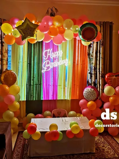 A unique and colorful fruit-themed birthday party. This vibrant setup includes a multi-colored fringe backdrop, fruit-shaped foil balloons, and a bright neon 'Happy Birthday' sign.