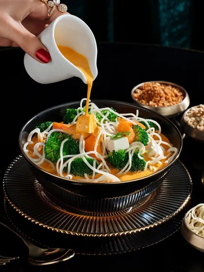A dynamic pour shot of a creamy broth into a bowl of Khowsuey at Club Jolies. The dark background makes the vibrant yellow of the broth and the fresh green broccoli pop, creating a very appetizing image.