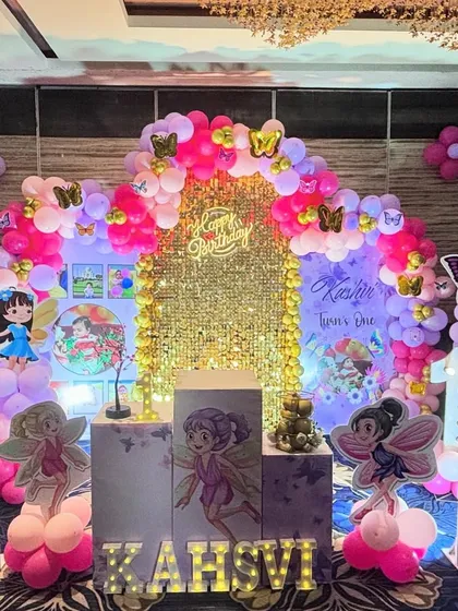 A full view of a fairy-themed birthday setup. The arrangement includes a shimmering gold backdrop, a multi-colored balloon arch with butterfly accents, and light-up letters spelling the child's name.