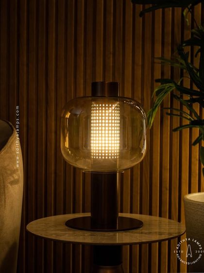 This table lamp features a perforated inner cylinder shielded by an outer amber glass shade. The design creates a mesmerizing pattern of light, offering a warm and intricate glow for a side table or console.