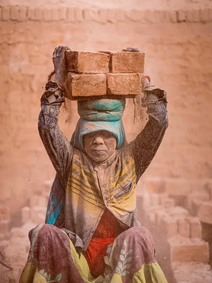 A woman, her face and clothes covered in dust, sits with a load of bricks on her head, her direct gaze telling a story of resilience.