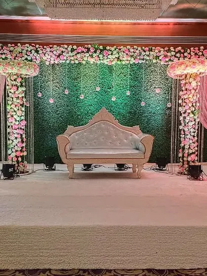 An extravagant reception stage with floral pillars, a lush green backdrop, and hanging light bulbs. The pink draping adds a soft, romantic touch to the overall grand design.