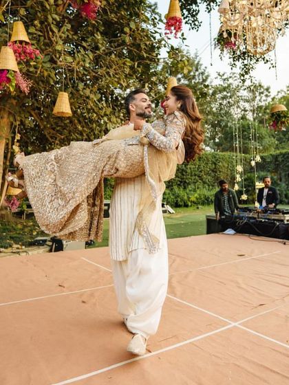 The groom sweeps his bride off her feet during their romantic couple's dance. This moment, set against a beautiful outdoor backdrop with hanging chandeliers, is what makes a Sangeet performance so special.