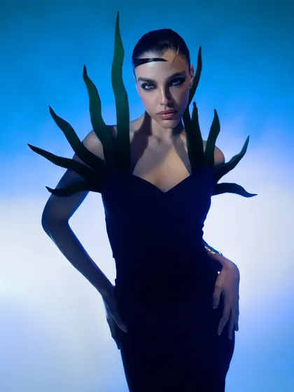 A conceptual fashion portrait with a striking silhouette. The green, plant-like shoulder pieces and the deep blue background create a surreal, almost alien-like visual.