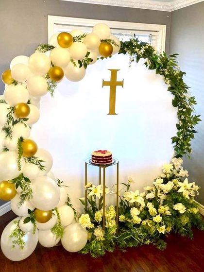 An elegant decoration for a First Holy Communion, combining white balloons with fresh greenery and white lilies. A golden cross is placed on the central backdrop.
