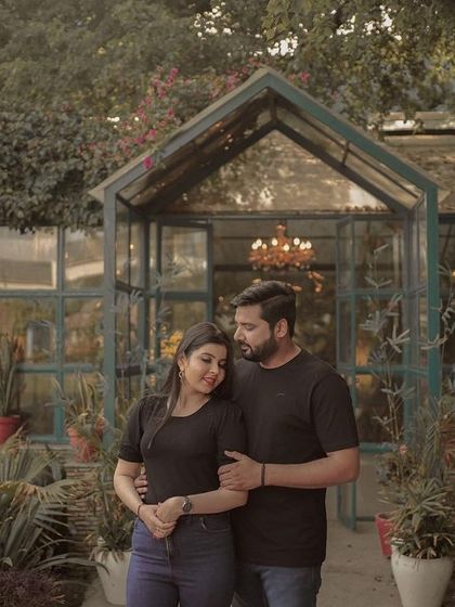 A romantic pre-wedding portrait of a couple in a lush garden, standing before a beautiful greenhouse, capturing a moment of quiet connection.