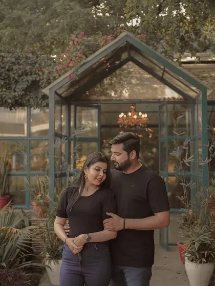 A romantic pre-wedding portrait of a couple in a lush garden, standing before a beautiful greenhouse, capturing a moment of quiet connection.