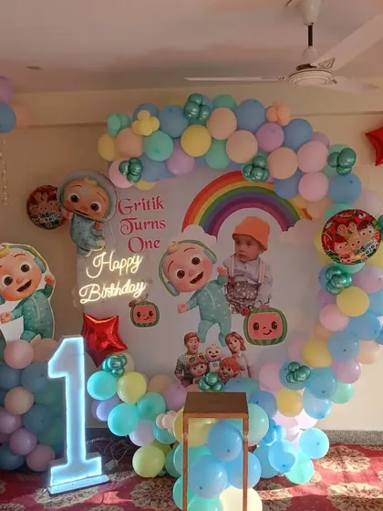 A Cocomelon themed first birthday party for "Gritik." This at-home setup features a circular balloon arch in pastel colors, a personalized backdrop with the baby's photo, and a light-up number "1."