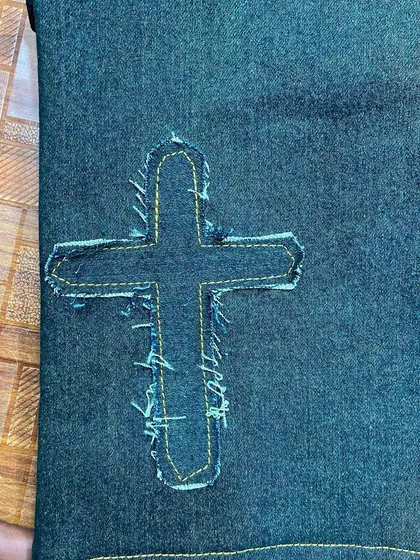 A close-up of the frayed cross patch detail. Notice the precise stitching that secures the patch while allowing for a rugged, textured edge.