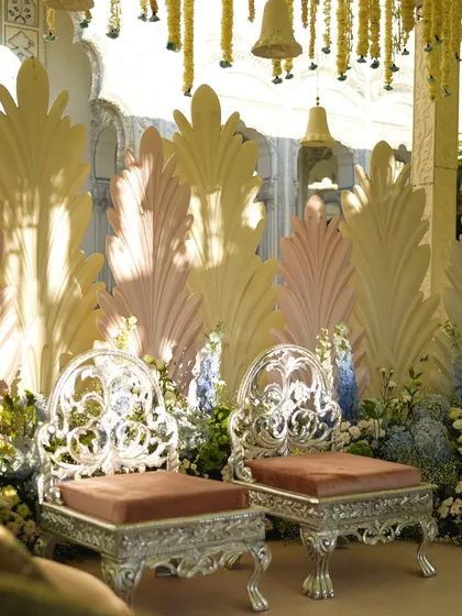 The elegant silver chairs for the couple at the mandap, set against the backdrop of pastel feather-like panels.