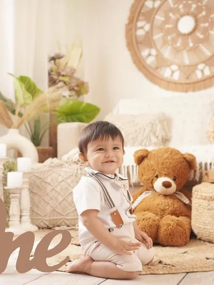 A charming first birthday portrait of a baby boy in a boho-chic setup, featuring a large wooden 'One' sign and a friendly teddy bear.